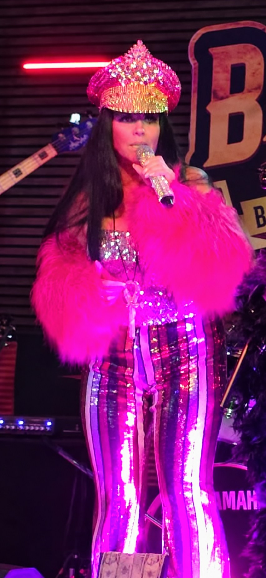 Sue Mondani singing live in a sparkling rhinestone hat, pink fur jacket and rainbow sequin pants